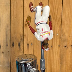 Peut inclure: Un pommeau de robinet sur le thème du baseball représentant un joueur de baseball en uniforme blanc avec des accents rouges, tenant un gant. La figurine est à l'envers. Le pommeau est fixé à un robinet chromé.