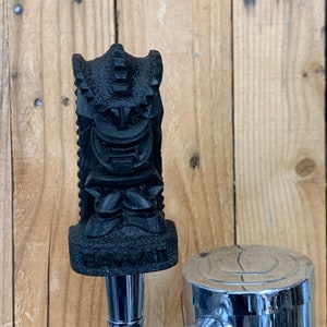May include: Black tiki-style beer tap handle with the word "Hawaii" on the bottom. The handle is attached to a silver beer tap.