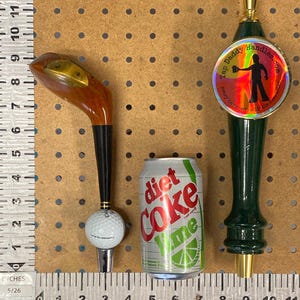 Puede incluir: Un surtido de art&iacute;culos que incluyen un tirador de grifo con forma de palo de golf, una lata de Diet Coke Lime y un tirador de grifo verde con el texto "Tap Daddy Handles.com". El tirador de grifo con forma de palo de golf tiene una cabeza de madera y una base de pelota de golf.
