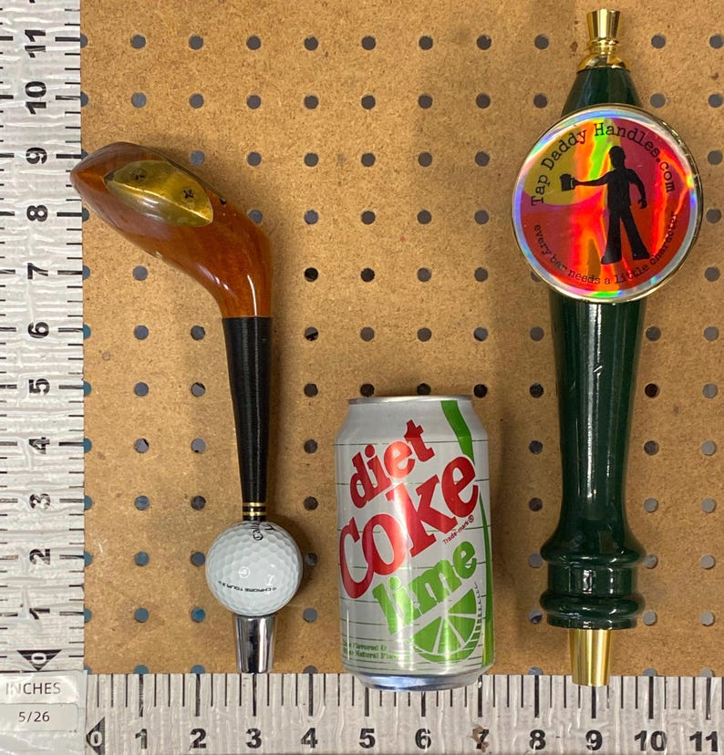 Puede incluir: Un surtido de art&iacute;culos que incluyen un tirador de grifo con forma de palo de golf, una lata de Diet Coke Lime y un tirador de grifo verde con el texto "Tap Daddy Handles.com". El tirador de grifo con forma de palo de golf tiene una cabeza de madera y una base de pelota de golf.