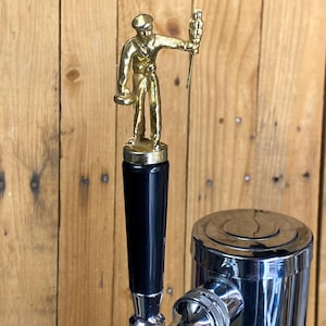 May include: A brass beer tap handle featuring a figure of a man holding a tray and a torch. The handle has a black, cylindrical grip and is attached to a chrome-finished tap. The background is a wooden wall.