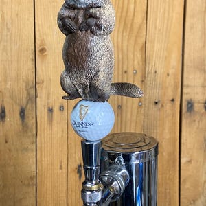 May include: A novelty beer tap handle featuring a groundhog figure atop a golf ball with the Guinness logo. The groundhog is brown and gray, and the tap is chrome.