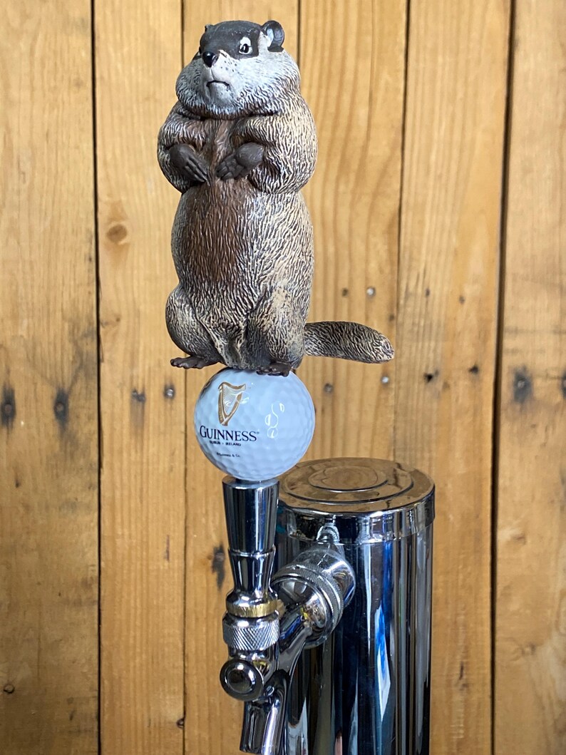 May include: A novelty beer tap handle featuring a groundhog figure atop a golf ball with the Guinness logo. The groundhog is brown and gray, and the tap is chrome.