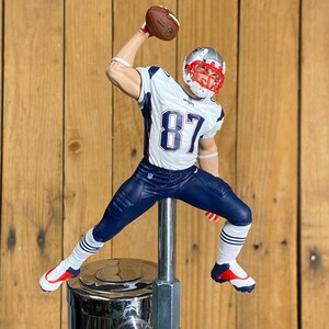 May include: A figurine of an American football player in a white, blue, and red uniform, throwing a brown football. The figure is mounted on a chrome tap handle. The player wears a helmet with a red face mask and the number 87.