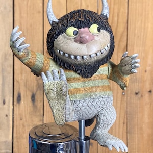 May include: A Where the Wild Things Are Max character tap handle. The figure has a tan face, yellow eyes, and a pink nose. It wears a striped orange and green shirt and has white claws. The tap handle is mounted on a chrome base.