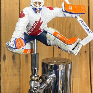 May include: A hockey goalie figurine wearing a white and red jersey with the word "CANADA" on the front, a blue and orange helmet, and a white and orange hockey stick. The figurine is mounted on a silver beer tap.