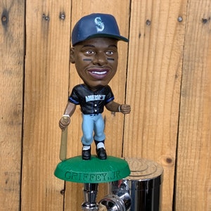 May include: A green and white baseball-themed beer tap handle featuring a bobblehead of a baseball player wearing a blue and white baseball cap with a white 'S' on it. The handle is inscribed with the name "GRIFFEY, JR."