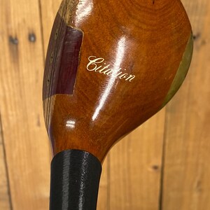 Puede incluir: Un palo de golf de madera vintage con una cabeza marr&oacute;n oscuro y un agarre negro. La cabeza del palo tiene la palabra "Citation" en cursiva. La veta de la madera es visible en la cabeza del palo.