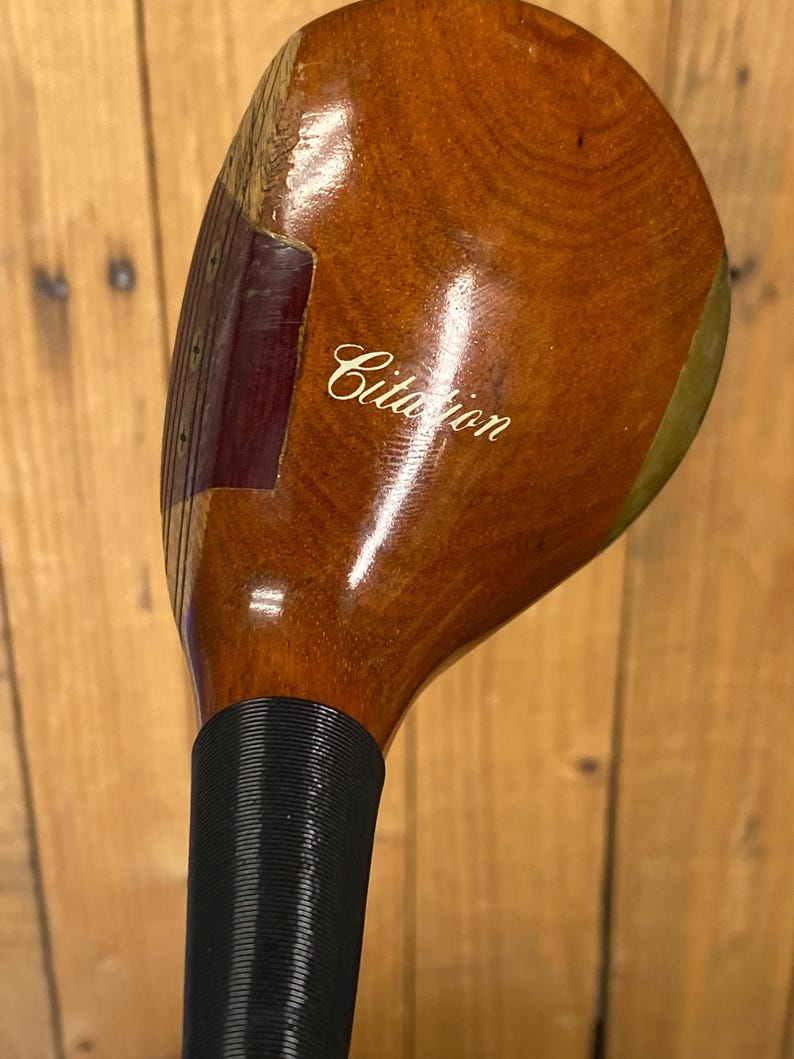 Puede incluir: Un palo de golf de madera vintage con una cabeza marr&oacute;n oscuro y un agarre negro. La cabeza del palo tiene la palabra "Citation" en cursiva. La veta de la madera es visible en la cabeza del palo.
