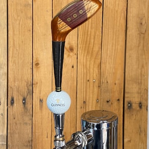 Puede incluir: Un grifo de cerveza personalizado con una cabeza de palo de golf y un tirador de grifo de bola de golf Guinness. El grifo es de madera, negro y metal plateado. El grifo est&aacute; montado sobre un fondo de madera.