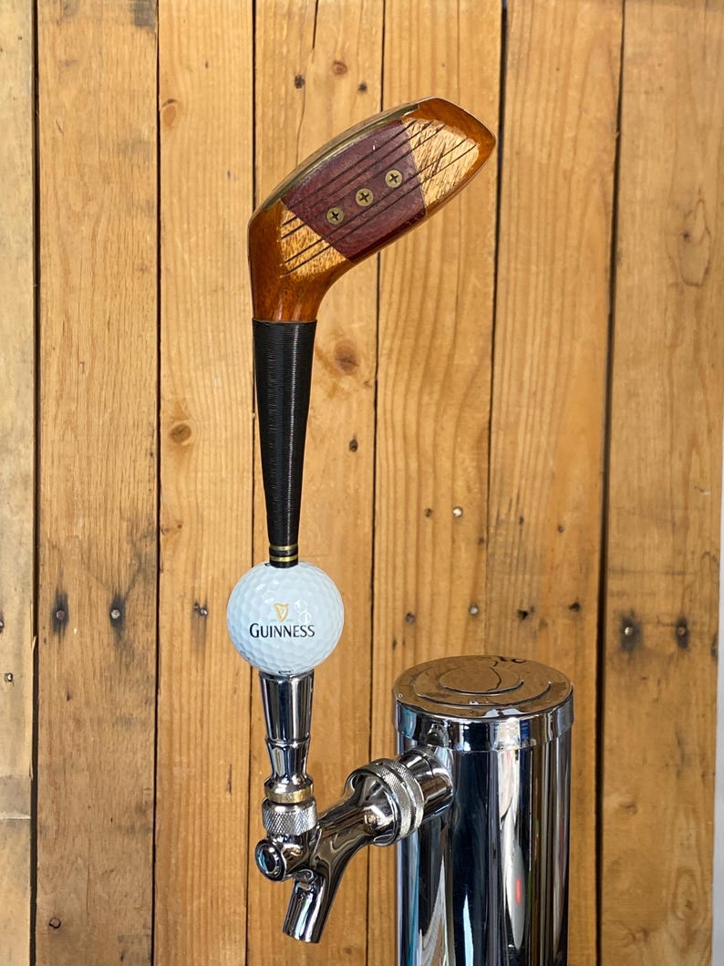 Puede incluir: Un grifo de cerveza personalizado con una cabeza de palo de golf y un tirador de grifo de bola de golf Guinness. El grifo es de madera, negro y metal plateado. El grifo est&aacute; montado sobre un fondo de madera.