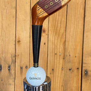 Puede incluir: Un tirador de grifo personalizado con una cabeza de palo de golf vintage. La cabeza del palo de madera es marr&oacute;n con un acento rojo, y el mango es negro. Una pelota de golf Guinness se encuentra encima de un grifo cromado.