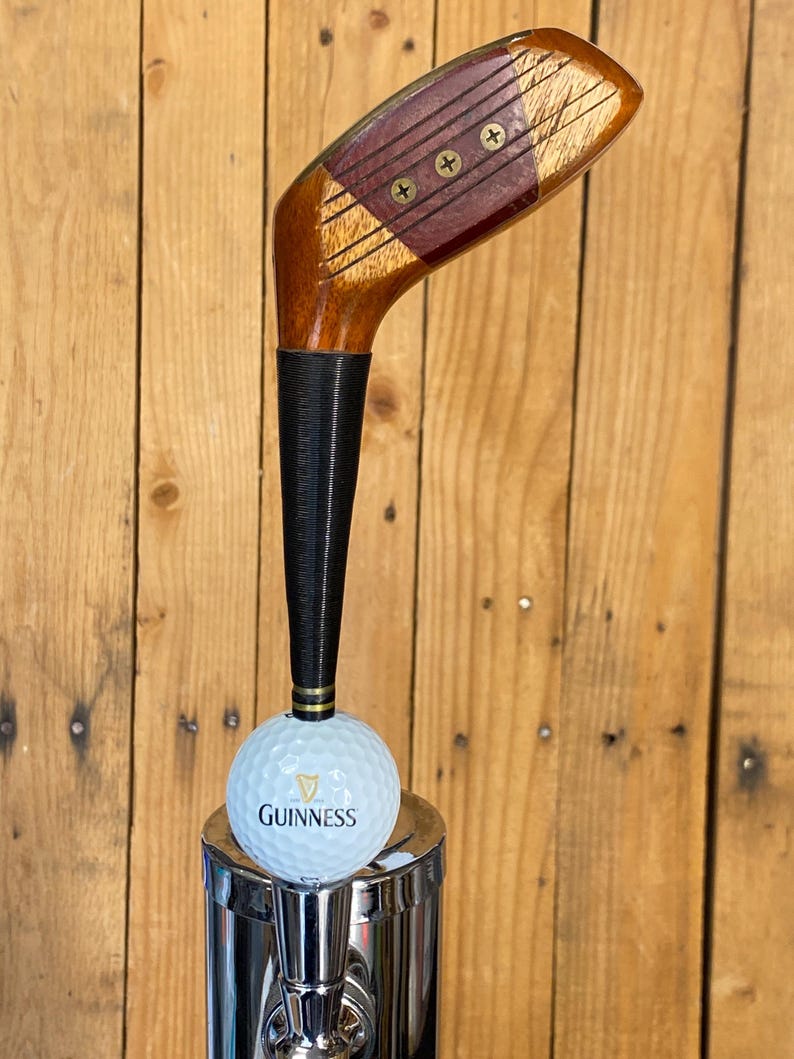 Puede incluir: Un tirador de grifo personalizado con una cabeza de palo de golf vintage. La cabeza del palo de madera es marr&oacute;n con un acento rojo, y el mango es negro. Una pelota de golf Guinness se encuentra encima de un grifo cromado.