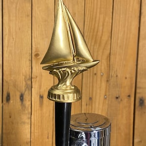 May include: A gold-colored sailboat tap handle with a black handle and chrome base. The sailboat is detailed with a mast and sails, mounted on a decorative base. The tap handle is designed for a beer or beverage dispenser.