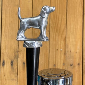 May include: A silver-colored dog figurine stands atop a black and chrome tap handle. The dog is a detailed sculpture, with its tail raised and head tilted upwards. The tap handle is part of a larger chrome and silver beer tap system.