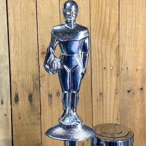 May include: A chrome-finished football tap handle featuring a stylized figure holding a football. The figure stands atop a circular base, with a cylindrical component and a lever visible below. The background is a wooden surface.