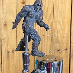 Peut inclure: Une figurine de Bigfoot brune avec une barbe, debout sur un robinet de bière chromé.