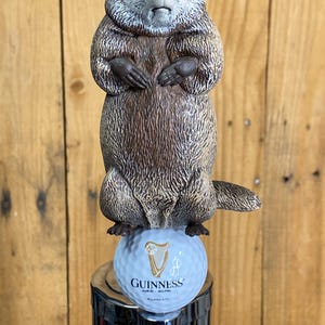 May include: A novelty tap handle featuring a groundhog perched atop a golf ball with the Guinness logo. The groundhog is brown and gray, with a detailed fur texture. The golf ball is light blue and the tap handle is chrome.