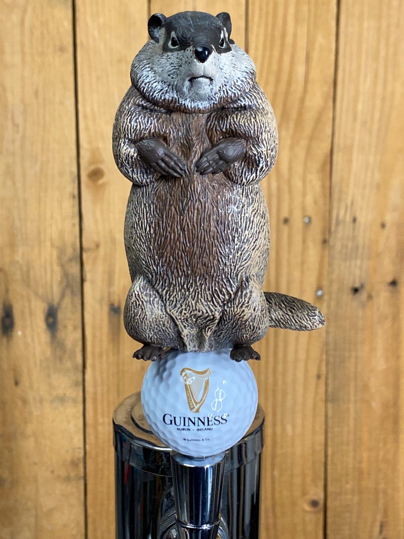 May include: A novelty tap handle featuring a groundhog perched atop a golf ball with the Guinness logo. The groundhog is brown and gray, with a detailed fur texture. The golf ball is light blue and the tap handle is chrome.