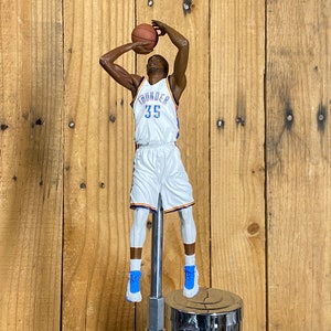 May include: A basketball-themed tap handle featuring a figurine of a basketball player in a white and blue uniform, mid-shot. The figure is mounted on a chrome tap. The background is a wooden wall.