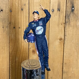 May include: A blue and white NASCAR driver figurine holding a helmet, standing on top of a chrome beer tap. The figurine is wearing a blue racing suit with the word "Rusty" on the chest.