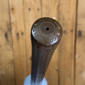 May include: A brown golf club grip with the word "CONQUEST" and "MADE IN U.S.A." printed on the end. The grip is textured with vertical lines and is attached to a white golf ball.