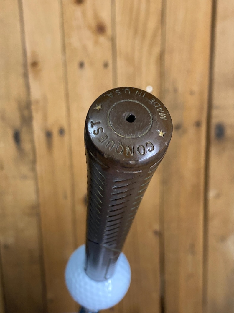 May include: A brown golf club grip with the word "CONQUEST" and "MADE IN U.S.A." printed on the end. The grip is textured with vertical lines and is attached to a white golf ball.