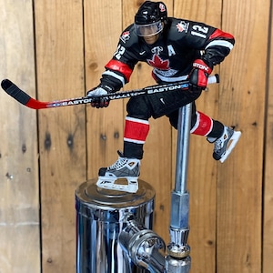 Peut inclure: Une figurine de joueur de hockey portant un maillot noir et rouge avec le mot "CANADA" sur le devant, tenant un bâton de hockey noir et blanc. La figurine est debout sur un robinet de bière argenté.
