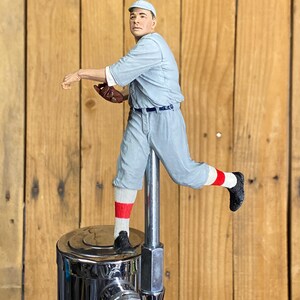 May include: A baseball-themed tap handle featuring a figurine of a baseball player in a throwing pose. The player is wearing a light blue uniform, a cap, and black shoes. The tap handle is made of chrome and is mounted on a wooden background.