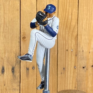 May include: A baseball player figurine wearing a white uniform with blue trim and a blue hat with a white 'S' on it. The figurine is holding a black baseball glove and is in a pitching pose. The figurine is mounted on a silver metal tap handle.