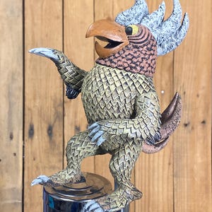 May include: A whimsical, hand-painted tap handle featuring a bird-like creature with a brown beak, patterned feathers, and a crest of blue and white feathers. The creature is posed with one arm outstretched, standing on a chrome tap.