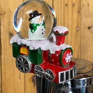 May include: A Christmas-themed snow globe featuring a snowman inside a clear glass sphere. The globe sits atop a red and green toy train with silver wheels. The train is decorated with white glitter, suggesting a snowy scene.