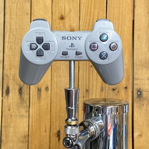 May include: A gray PlayStation controller is mounted on a chrome beer tap. The controller has the words "SONY" and "PlayStation" printed on it. The controller is attached to the tap with a metal rod.