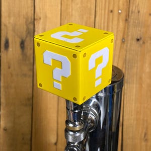 May include: A yellow cube-shaped tap handle featuring white question marks on each face. The tap handle is mounted on a chrome faucet. The background is wooden. The design is inspired by a video game.