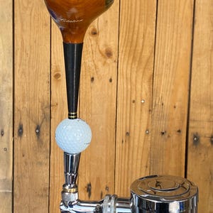 Puede incluir: Un tirador de grifo de cerveza personalizado con una cabeza de palo de golf, una pelota de golf y un grifo cromado. La cabeza del palo de golf es marr&oacute;n con detalles negros. La pelota de golf es blanca. El grifo es cromado.