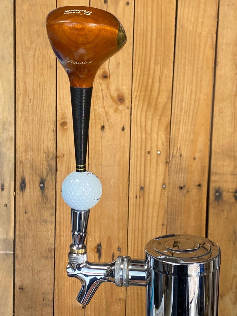 Puede incluir: Un tirador de grifo de cerveza personalizado con una cabeza de palo de golf, una pelota de golf y un grifo cromado. La cabeza del palo de golf es marr&oacute;n con detalles negros. La pelota de golf es blanca. El grifo es cromado.