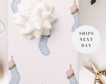 May include: White wrapping paper with a repeating pattern of blue and pink Christmas stockings. A large white bow sits on top of the paper. Gold scissors and the text "SHIPS NEXT DAY" are also visible.