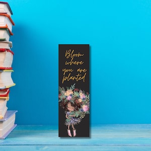 May include: Black bookmark with a gold inscription that reads "Bloom where you are planted". A cartoon girl with brown hair is holding a bouquet of flowers.