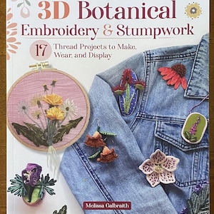 May include: A book titled "3D Botanical Embroidery & Stumpwork" with examples of embroidered floral designs. The book cover features a denim jacket adorned with various 3D embroidered flowers and a framed embroidery hoop with yellow flowers.
