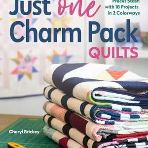 May include: A stack of colorful quilts with a book titled "Just One Charm Pack Quilts" and a rotary cutter. The quilts feature various patterns and colors, including blue, red, and white. The book cover also has text that reads "BUST Your Precut Stash with 18 Projects in 2 Colorways."
