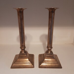 Può includere: Un paio di candelabri antichi in ottone con basi quadrate e colonne scanalate. I candelabri sono in buone condizioni con alcuni segni di età e usura.