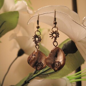 May include: A pair of handcrafted earrings featuring brown shell pendants with copper wire accents. The earrings have a spiral design and are suspended from copper-colored hooks and decorative wire elements. The background includes white flowers and green leaves.