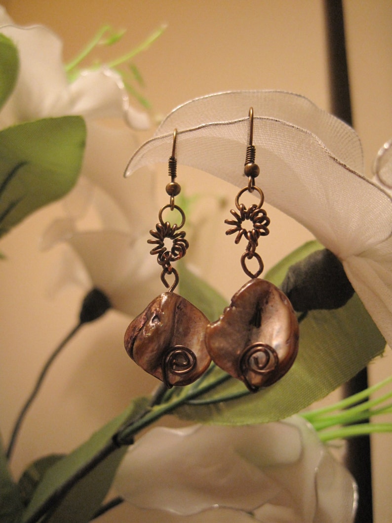 May include: A pair of handcrafted earrings featuring brown shell pendants with copper wire accents. The earrings have a spiral design and are suspended from copper-colored hooks and decorative wire elements. The background includes white flowers and green leaves.
