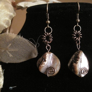 May include: A pair of handcrafted earrings featuring iridescent, pearlescent shells. The earrings have bronze-colored wire accents, including spiral details and floral-shaped connectors. The hooks are a darker bronze color. The earrings are displayed on a black surface.