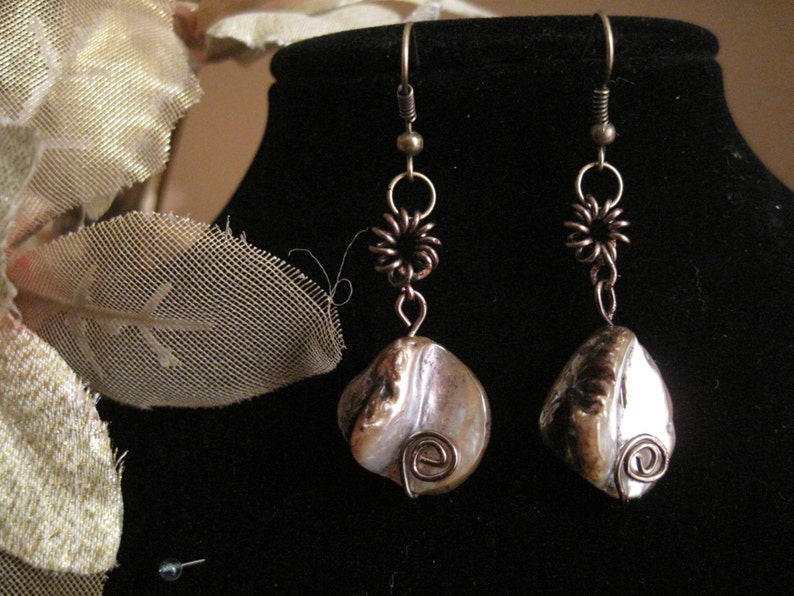 May include: A pair of handcrafted earrings featuring iridescent, pearlescent shells. The earrings have bronze-colored wire accents, including spiral details and floral-shaped connectors. The hooks are a darker bronze color. The earrings are displayed on a black surface.