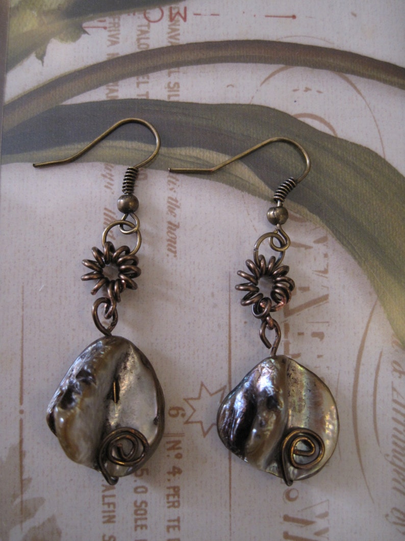 May include: A pair of handcrafted earrings featuring iridescent, shell-shaped pendants. The earrings have bronze-colored hooks, spiral wire accents, and small, flower-shaped embellishments. The earrings are designed for pierced ears.