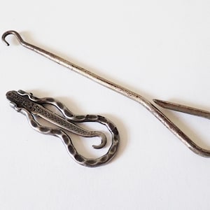 May include: Two antique metal tools on a white surface. One is a long, slender hook with a curved end. The other is a decorative, serpentine-shaped money clip. Both items have a tarnished, aged appearance.