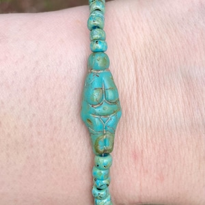 May include: Turquoise beaded bracelet featuring a central figurine of a stylized female form. The bracelet is strung with small, round turquoise beads, and the central figure has carved details.