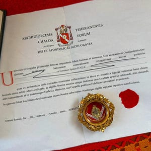 Pode incluir: Um documento com texto em latim, um selo de cera vermelho e um relicário com moldura dourada. O documento tem um brasão e está assinado. O relicário contém um pequeno objeto ornamentado com um design vermelho e dourado.