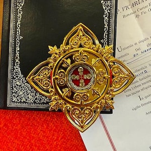 Pode incluir: Relíquia religiosa dourada ornamentada com um design central vermelho e prateado, colocada sobre um livro preto com bordas decorativas e um fundo vermelho. A relíquia tem um design de cruz.